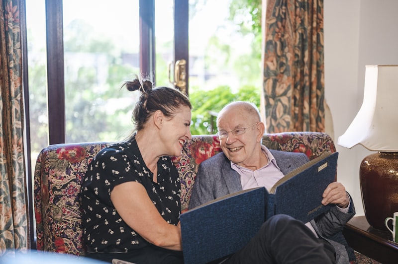 A companion caregiver and elderly man laughing together over a photo album in a warm home setting